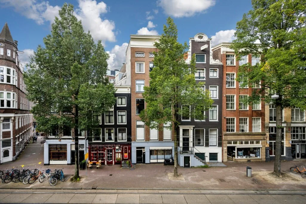 Gevels van historische panden aan de Nieuwezijds Voorburgwal in Amsterdam met bomen, fietsen en een restaurant op straatniveau.